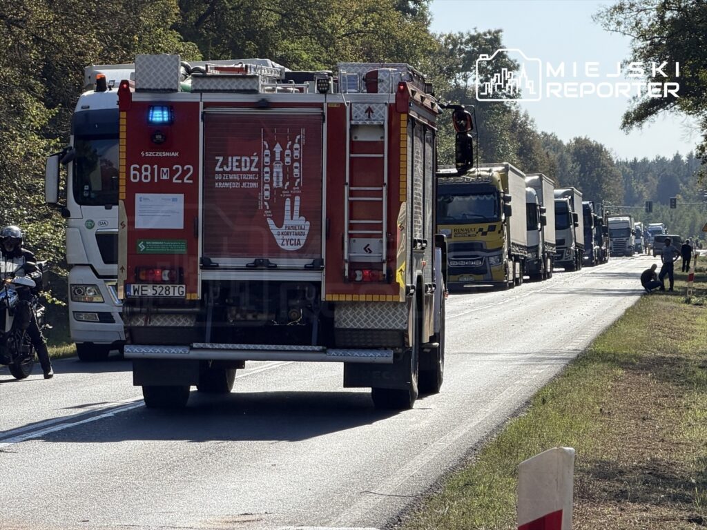 Czerwony wóz strażacki z włączonymi sygnalizatorami na drodze, obok długie kolejki ciężarówek i motocyklista.