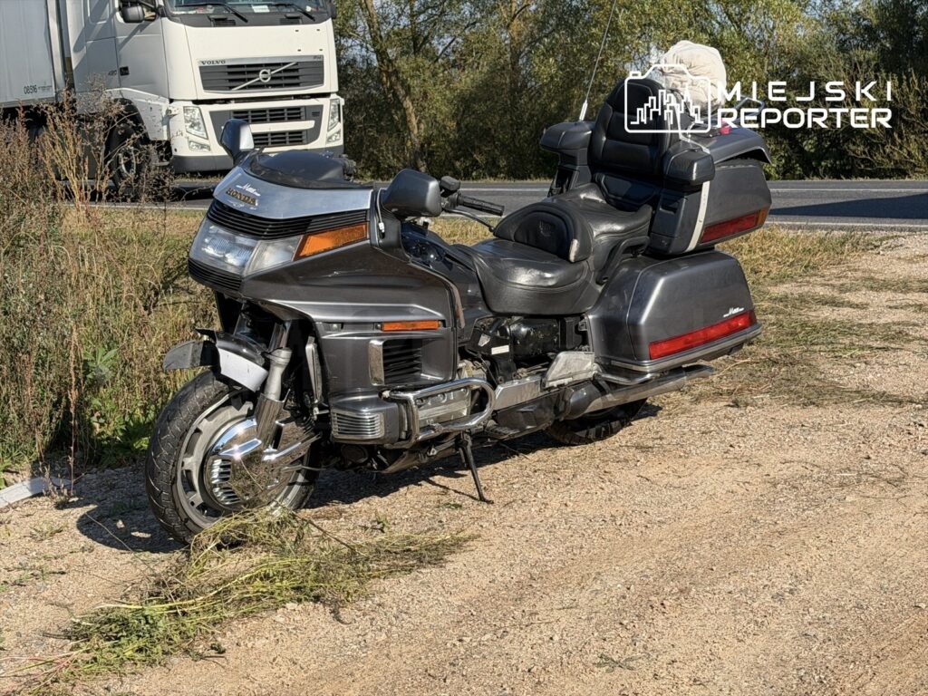 Uszkodzony motocykl leży na poboczu drogi, obok rosnących chwastów, w tle widoczny ciężarówka.