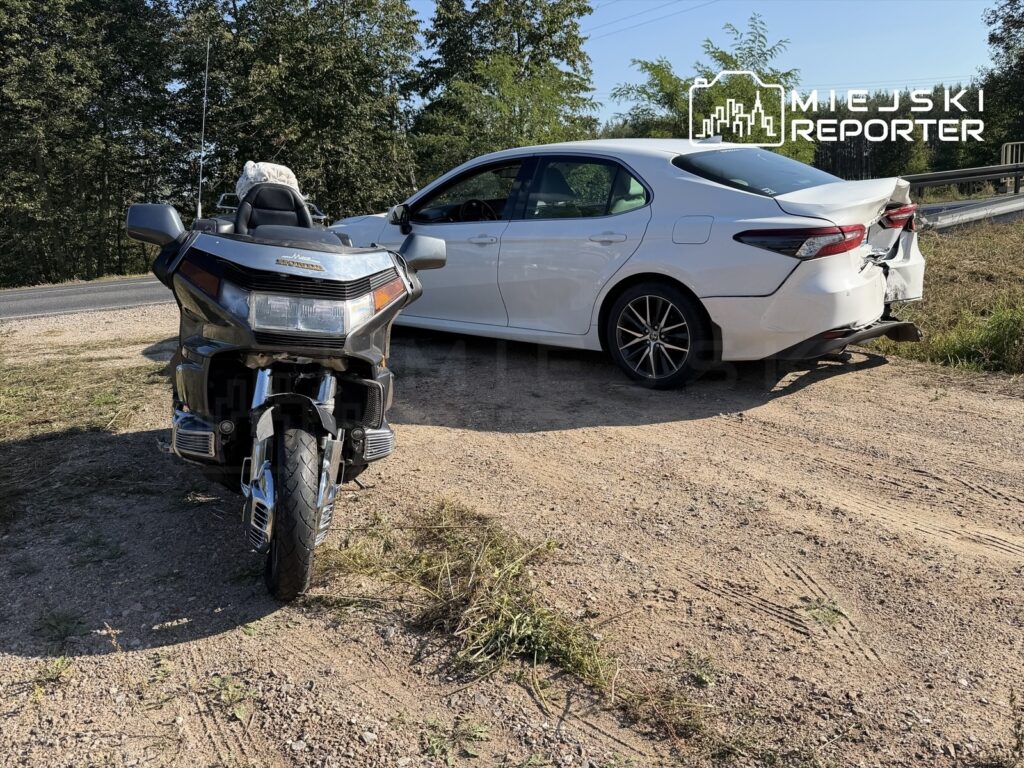 Motocykl stoi na poboczu drogi obok uszkodzonego białego samochodu osobowego.