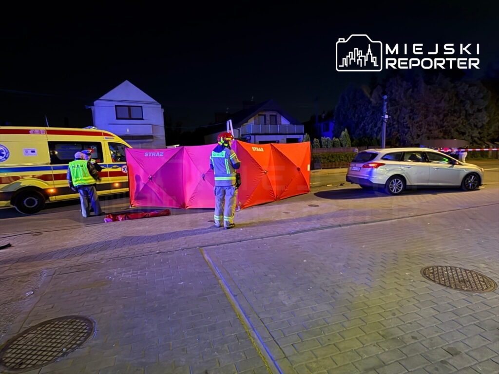 Ratownicy i policjanci pracują na miejscu wypadku, obok czerwonej osłony, na oświetlonej ulicy nocą.