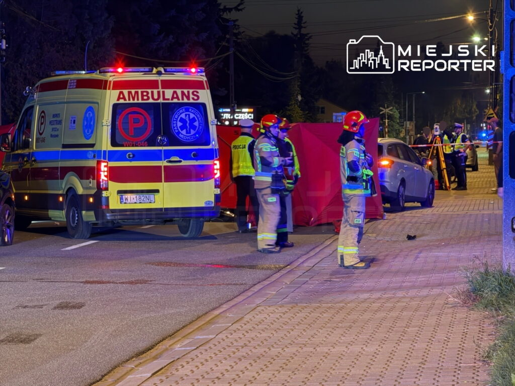Zespół ratunkowy i policja zabezpieczają miejsce wypadku drogowego, w tle widoczna karetka i czerwony namiot.