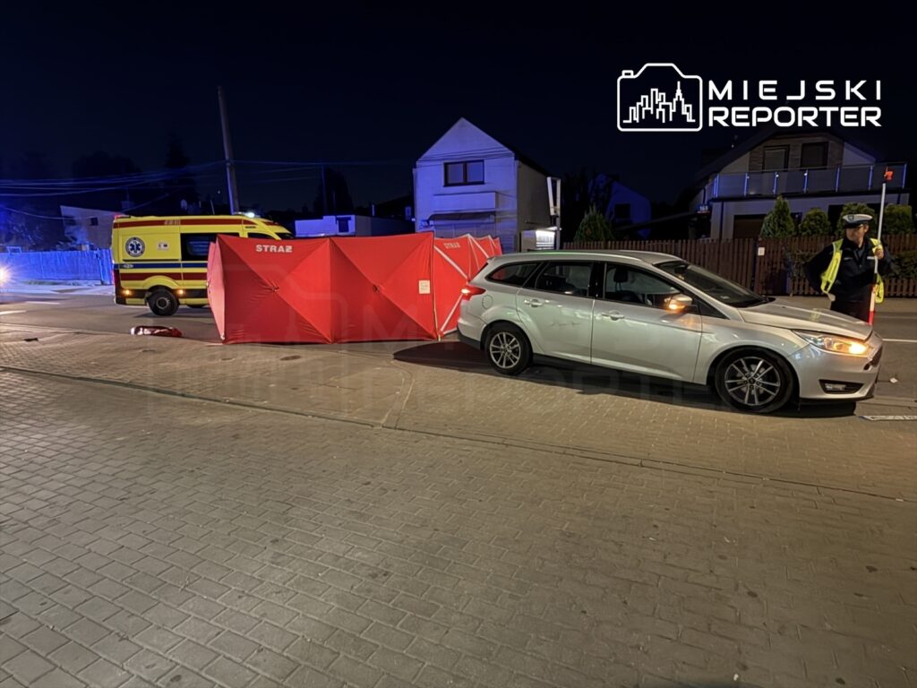 Na drodze stoi srebrny samochód osobowy, obok którego znajduje się czerwona osłona zabezpieczająca. W tle widać ambulans i funkcjonariusza policji kierującego ruchem.