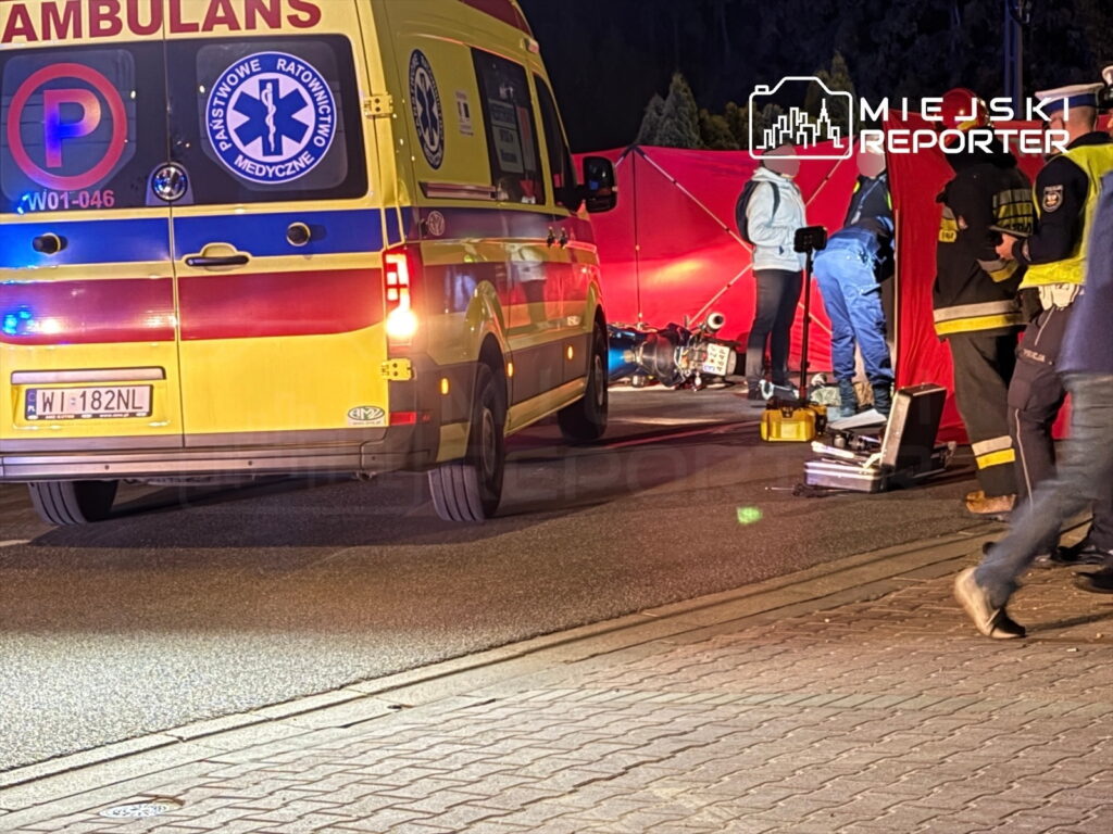 Ambulans z włączonymi sygnałami świetlnymi stoi na drodze, obok grupy ratowników medycznych i strażaków, zabezpieczających miejsce wypadku.