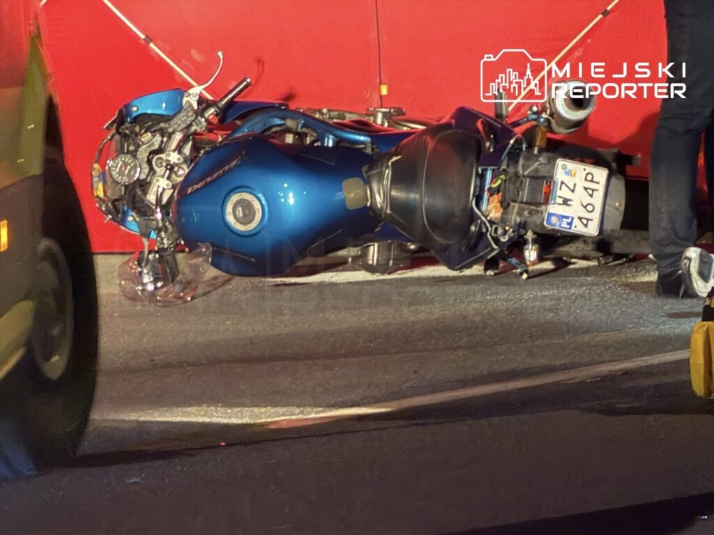 Motocykl leżący na drodze obok czerwonego pojazdu, z widocznymi uszkodzeniami, w otoczeniu służb ratunkowych.
