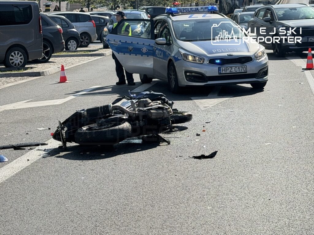 Motocykl leży na jezdni obok radiowozu policyjnego, w tle stoją zaparkowane samochody.