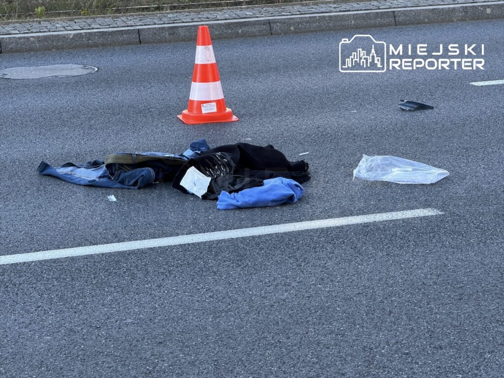 Na jezdni leżą porozrzucane ubrania oraz plastikowa torba, obok stoi pomarańczowy pachołek.
