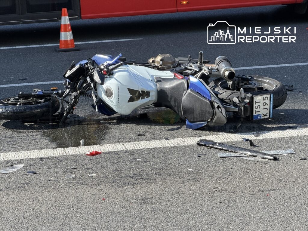 Motocykl leżący na jezdni z uszkodzoną karoserią, otoczony kawałkami szkła i plamą płynu. W tle widoczny autobus.