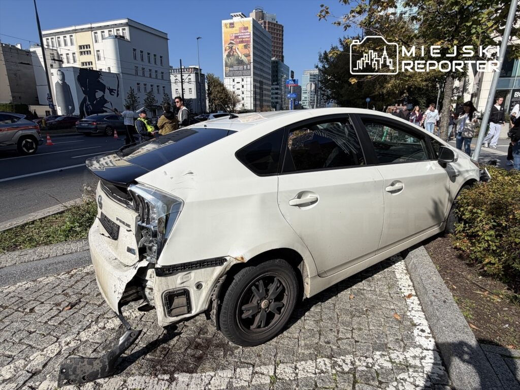 Biały samochód osobowy z uszkodzeniami, stojący na chodniku obok grupy osób rozmawiających z policjantem.