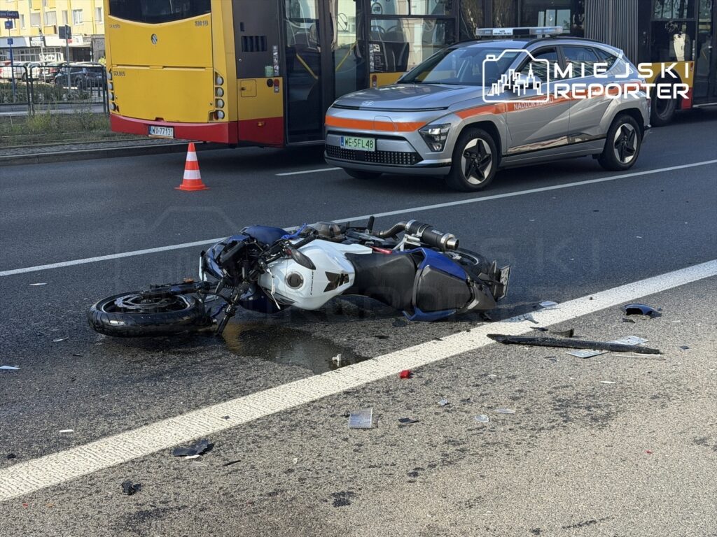 Motocykl leżący na jezdni, obok niego rozrzucone fragmenty pojazdu; w tle przejeżdżający samochód i autobus.