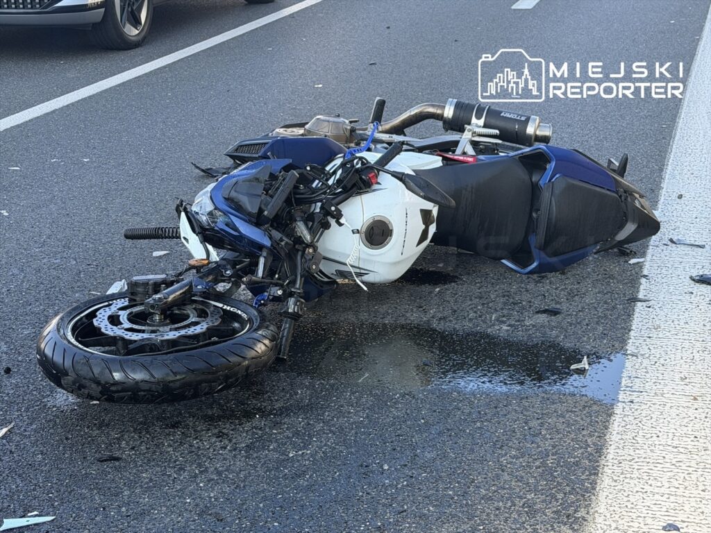 Motocykl leżący na drodze, z uszkodzoną przednią częścią i wyciekającymi płynami, obok przejeżdżające samochody.