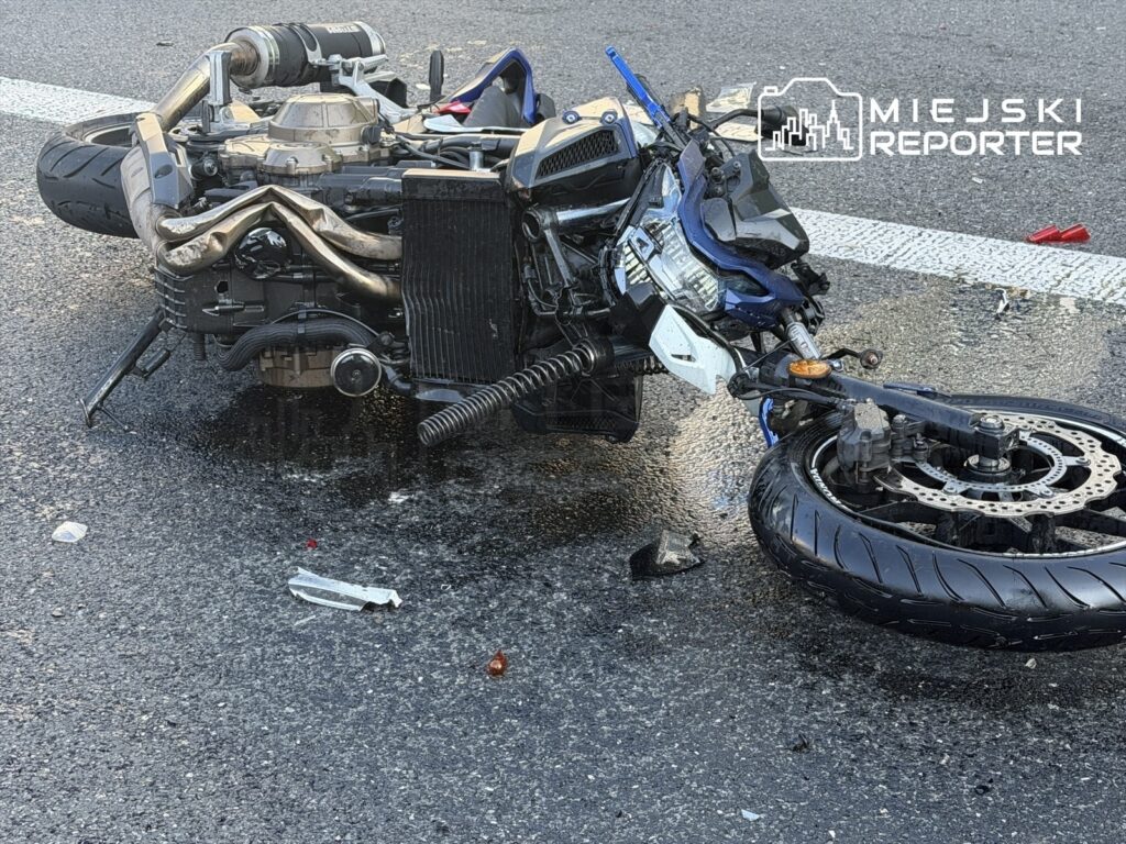 Motocykl leżący na boku na drodze, z uszkodzeniami silnika i odciekającymi płynami. W tle widać fragmenty rozbitego szkła.