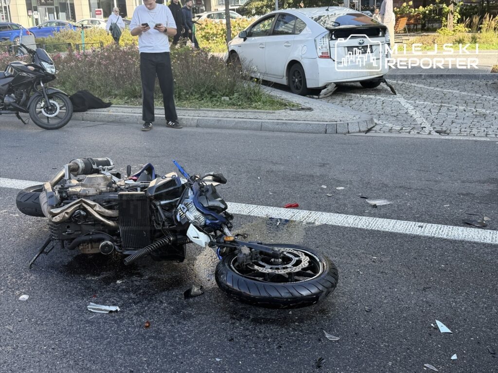 Motocykl leżący na boku na jezdni, w tle mężczyzna korzystający z telefonu, obok uszkodzony samochód.