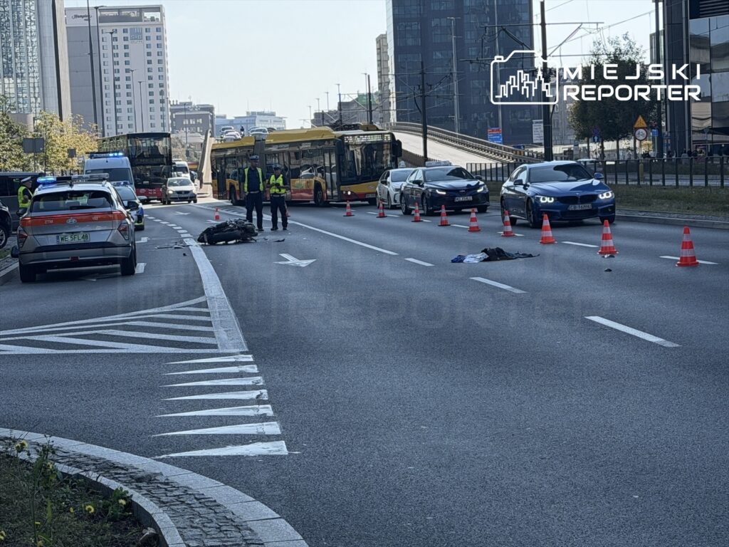 Na jezdni widać policjantów zabezpieczających miejsce wypadku, obok leży motocykl i ubrania. Ruch samochodowy jest zablokowany.