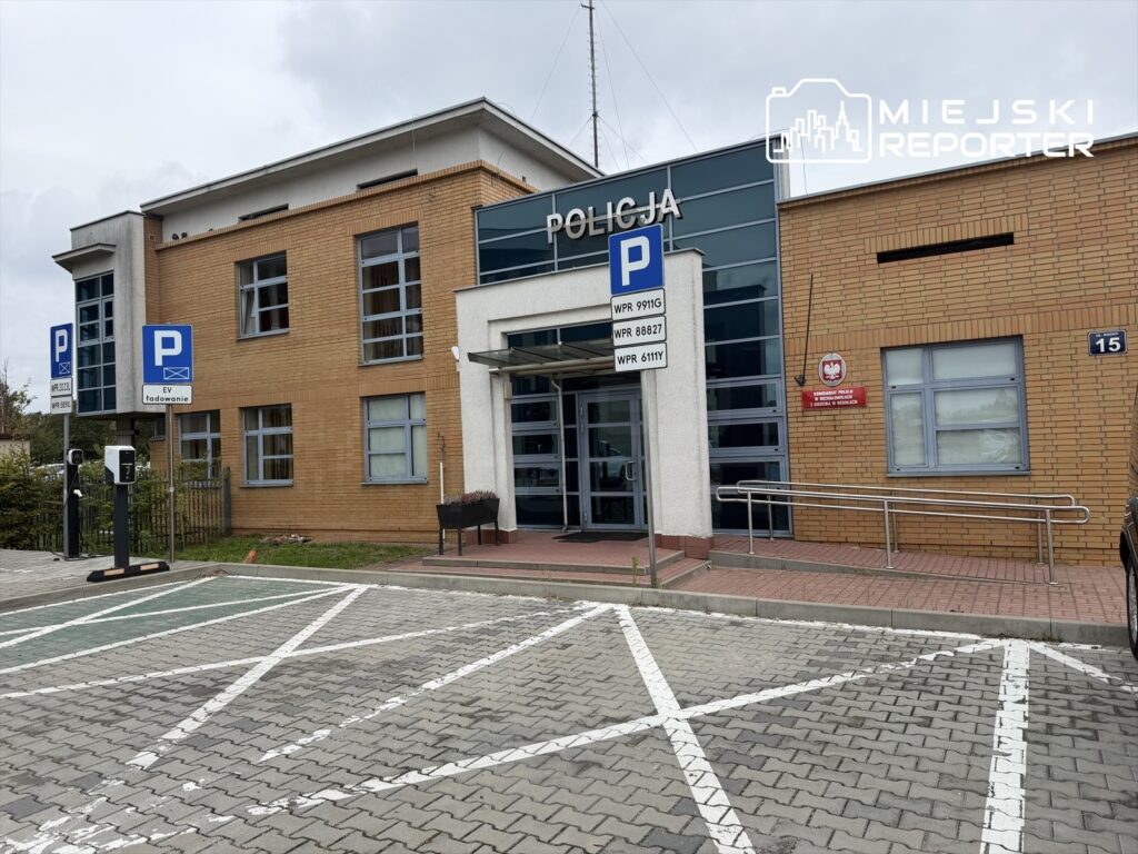 Budynek komisariatu policji z napisem „POLICJA” nad wejściem i parkingiem dla radiowozów przed wejściem.