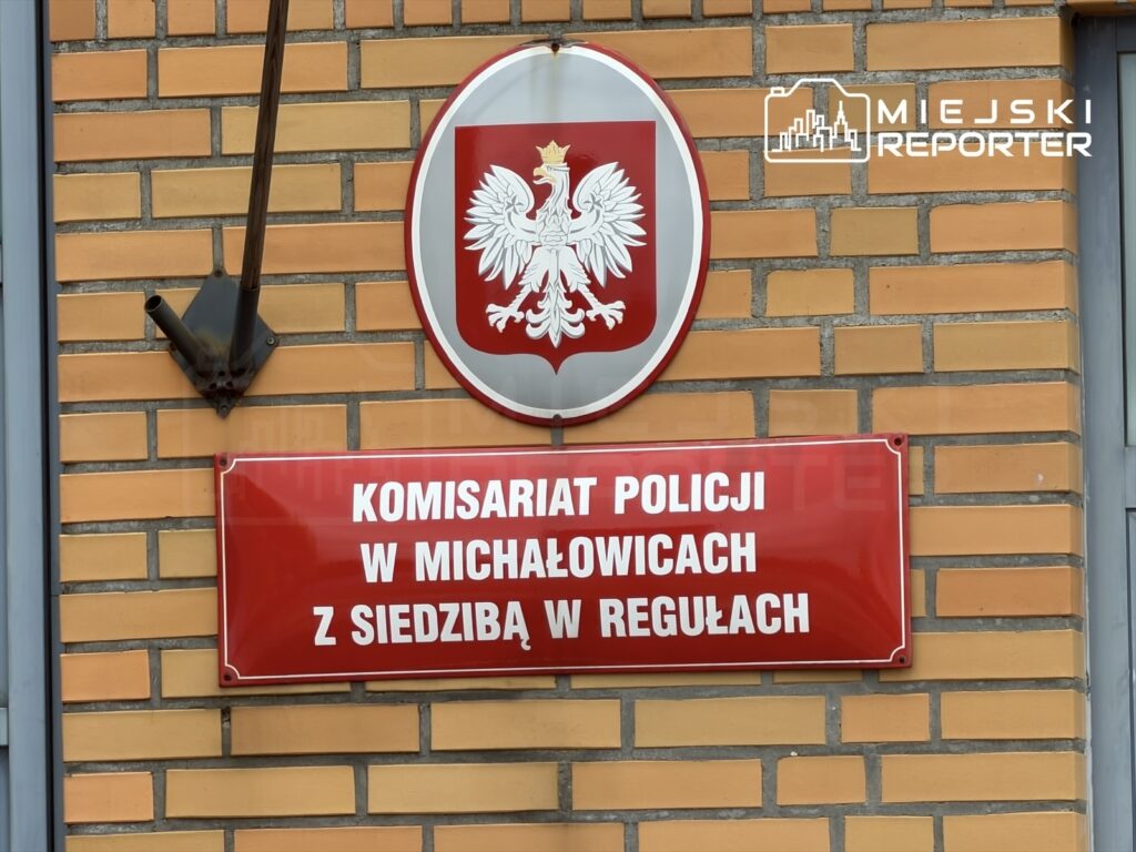 Czerwona tablica z napisem „Komisariat Policji w Michałowicach z siedzibą w Regułach” na ceglanej ścianie poniżej godła