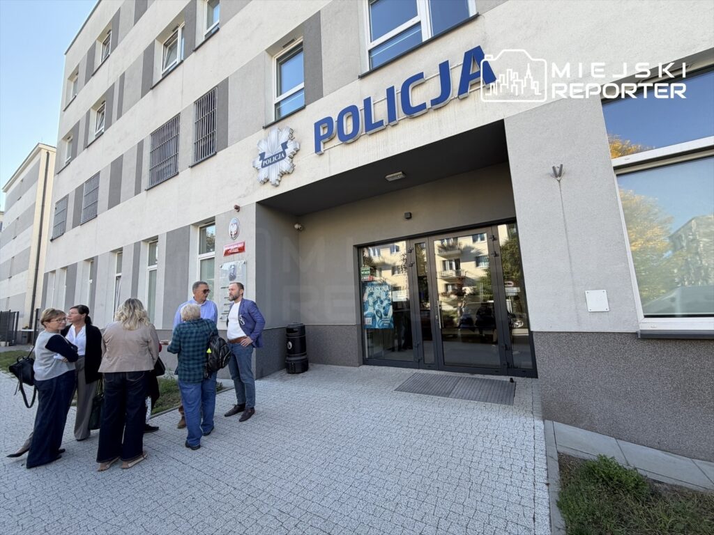 Grupa osób rozmawia przed wejściem do budynku z napisem „POLICJA”.
