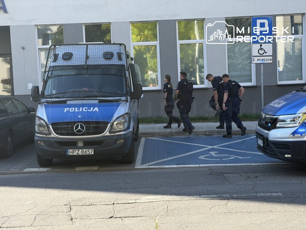 Policjanci idą wzdłuż budynku za zaparkowanym radiowozem, obok miejsca parkingowego dla osób z niepełnosprawnościami.