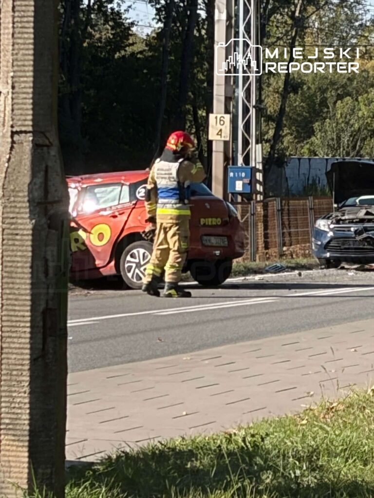 Strażak w hełmie stoi obok czerwonego samochodu, a przed nimi stoi uszkodzony samochód osobowy.