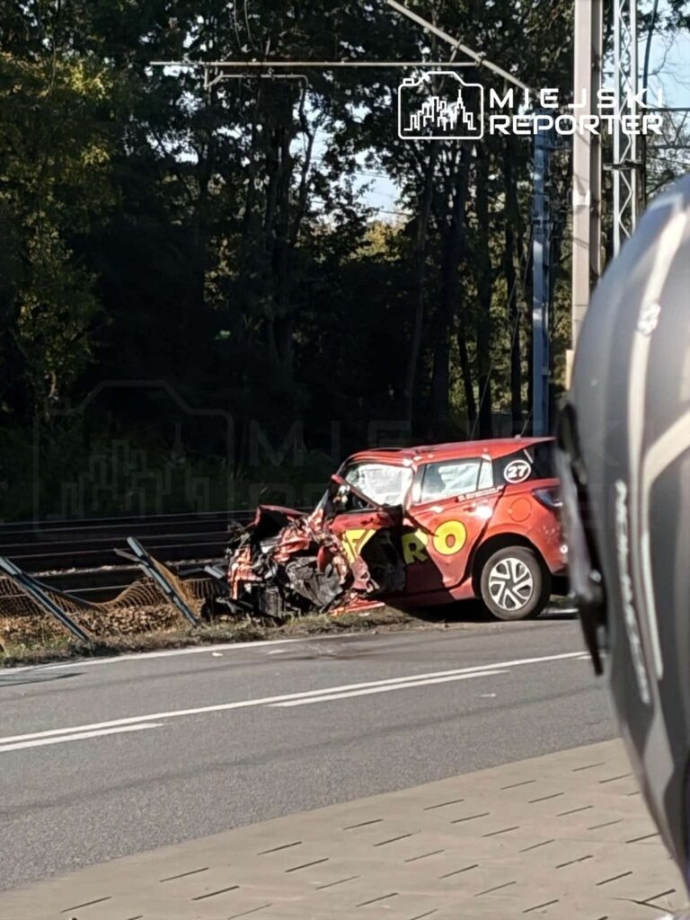 Czerwony samochód z mocno uszkodzonym przodem stoi przy drodze, obok przewróconego fragmentu ogrodzenia.