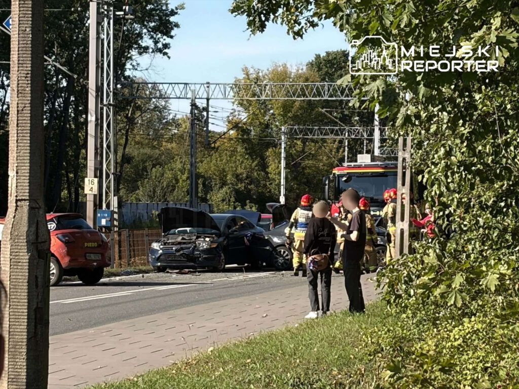 Uszkodzone samochody po zderzeniu na drodze, obok strażacy i osoby postronne; w tle wóz strażacki.