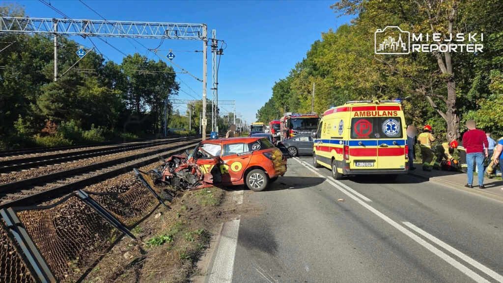Rozbity pomarańczowy samochód przy torach kolejowych, obok karetka pogotowia i służby ratunkowe na drodze.
