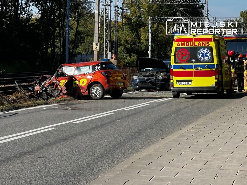 Rozbite czerwone auto i czarny samochód stoją na jezdni obok karetki pogotowia i torów kolejowych.