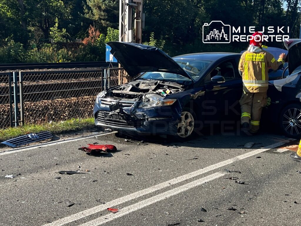 Strażak w żółtym ubraniu ochronnym stoi obok uszkodzonego samochodu z otwartą maską; na jezdni leżą odłamki.