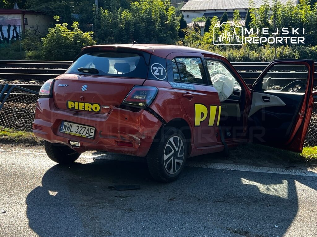 Uszkodzony czerwony samochód z otwartymi drzwiami i wystrzeloną poduszką powietrzną stoi przy torach kolejowych.