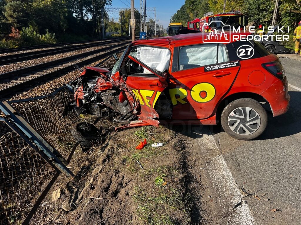 Czerwony samochód z mocno zniszczonym przodem stoi przy torach kolejowych, obok wykolejonego elementu infrastruktury torowej.