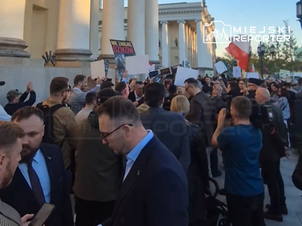 Tłum protestujących z transparentami i czerwonymi flagami przed budynkiem z kolumnami; na plakacie napis „NIE ZABIJAĆ DZIKÓW