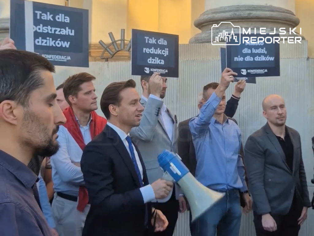 Mężczyzna w garniturze trzyma megafon wśród osób z tablicami „Tak dla redukcji dzika” i „Nie dla dzików”.