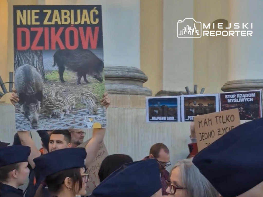 Uczestnicy protestu trzymają plakaty z hasłami „NIE ZABIJAĆ DZIKÓW” i „STOP RZĄDOM MYŚLIWYCH” w tłumie pod budynkiem.