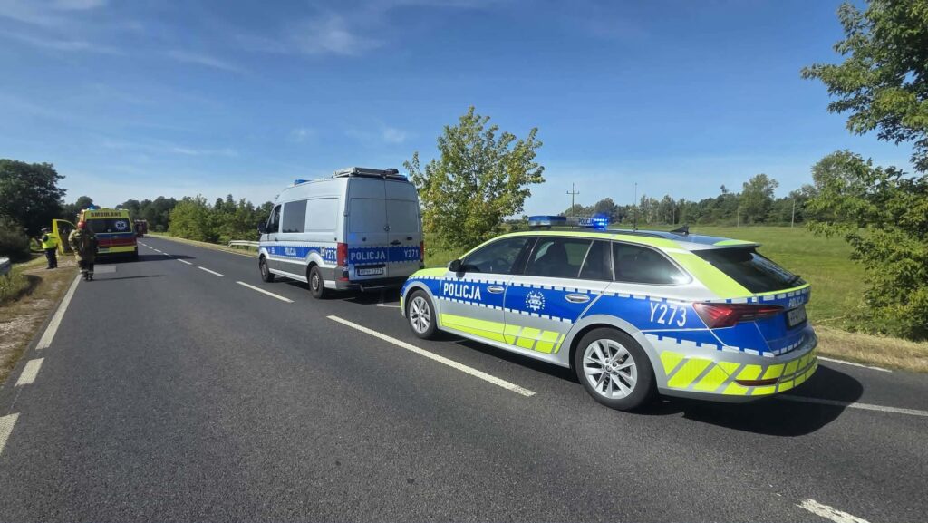 Na drodze stoją dwa radiowozy policyjne oraz karetka pogotowia. W tle widoczne są drzewa i błękitne niebo.