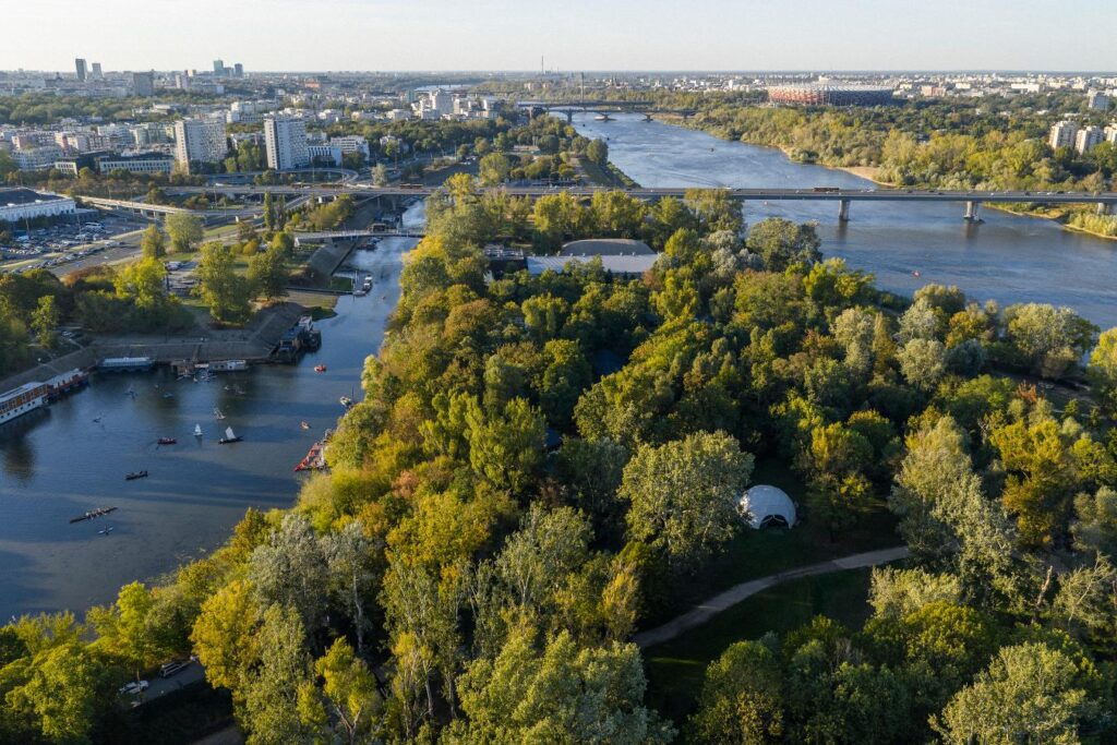 Widok na rzekę otoczoną zielonymi drzewami, w tle nowoczesne budynki i most, a na wodzie widać łodzie.