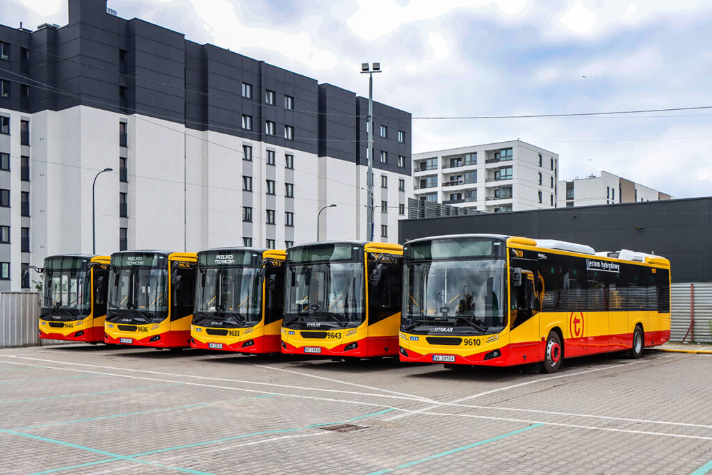 Pięć nowoczesnych autobusów hybrydowych zaparkowanych na terenie z płytkami, w tle budynki mieszkalne.