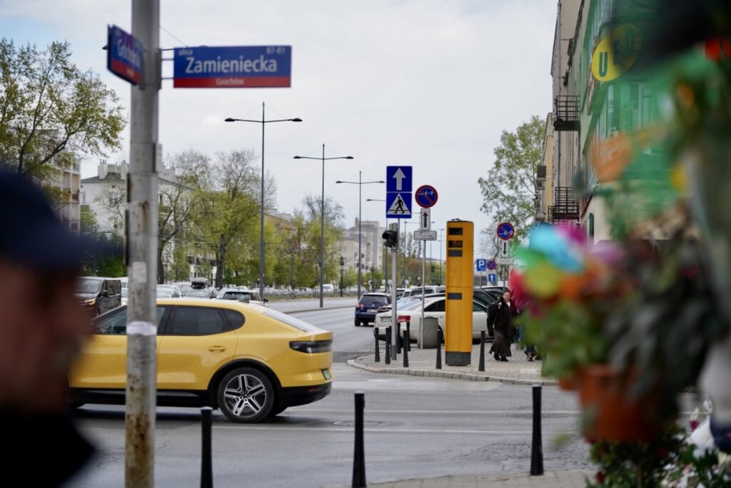 Żółty samochód osobowy zatrzymuje się na skrzyżowaniu, w tle widoczne znaki drogowe i ulica z drzewami.