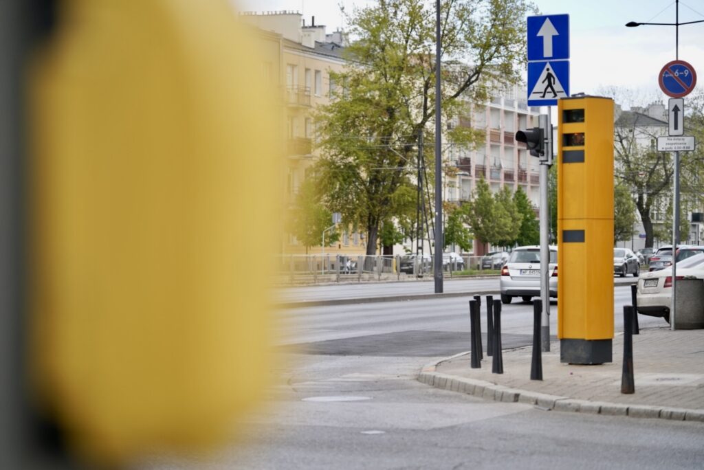 Żółty fotoradar ustawiony na skrzyżowaniu, w tle ruch uliczny oraz drzewa i budynki mieszkalne.
