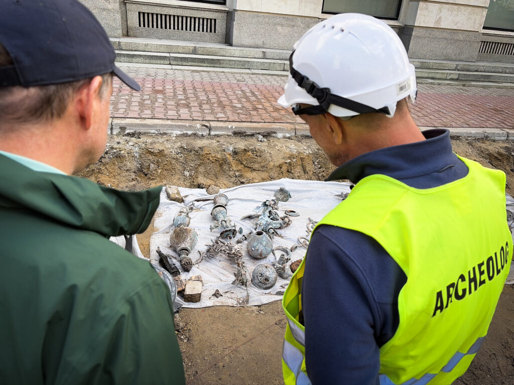 Dwaj mężczyźni w odzieży roboczej przyglądają się odkrytym artefaktom archeologicznym na wykopie.