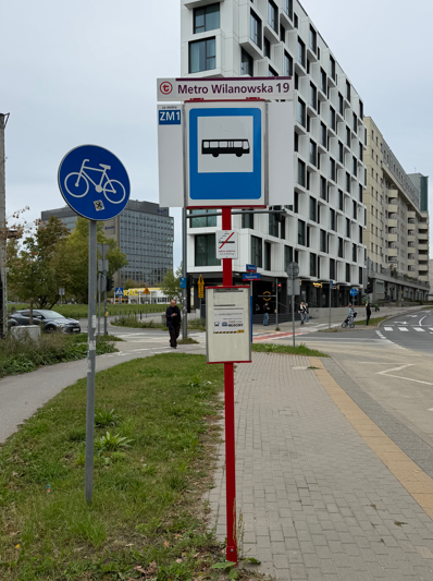 Przystanek autobusowy z oznaczeniem "Metro Wilanowska 19" w nowoczesnej zabudowie, obok znaku drogowym dla rowerzystów.