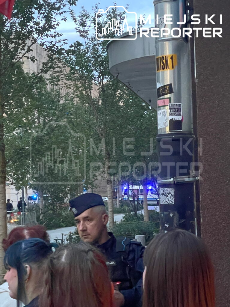 Funkcjonariusz policji rozmawia z grupą ludzi na ulicy, w tle widać niebieskie światła radiowozów, związane z ewakuacją.