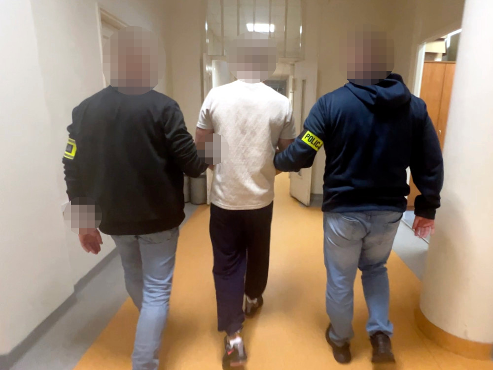Mężczyzna w jasnej koszulce prowadzony przez dwóch policjantów korytarzem budynku.