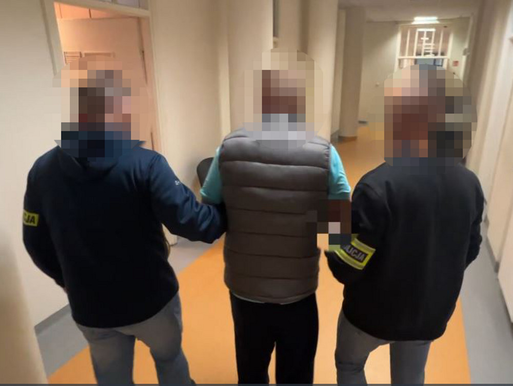 Trzech policjantów eskortuje mężczyznę w kamizelce, przechodząc przez korytarz w budynku.