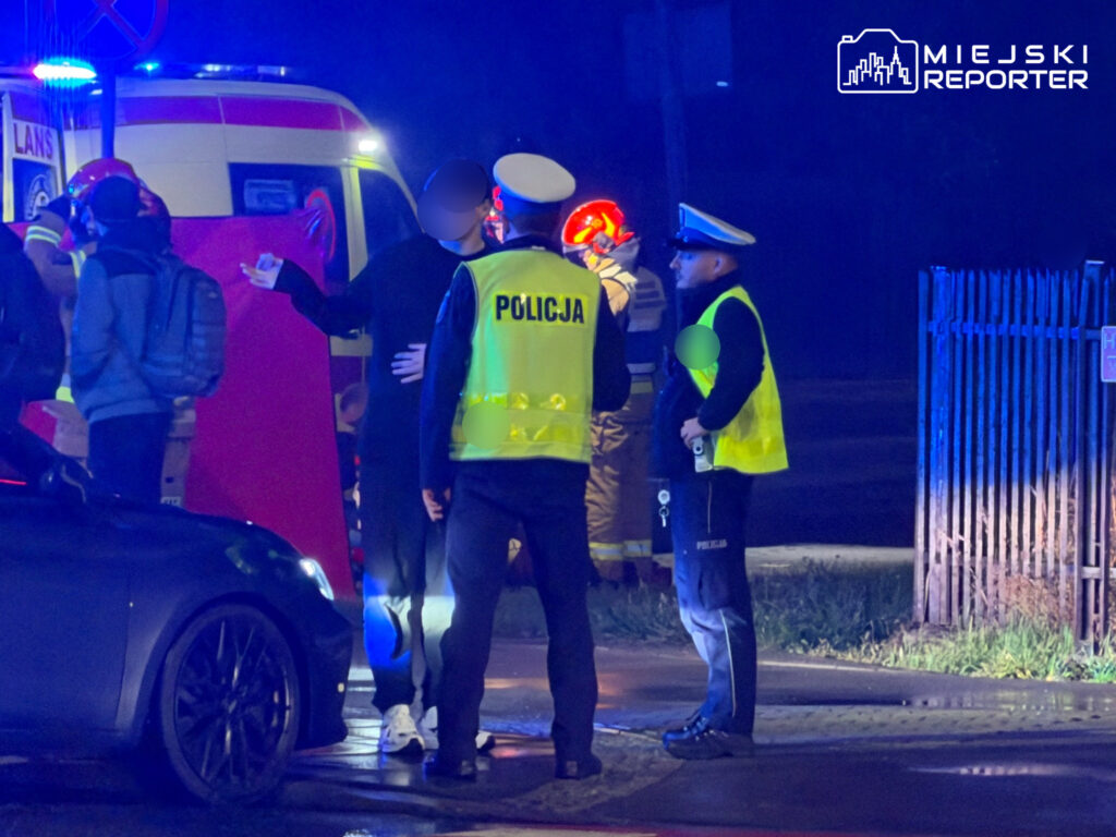 Policjanci w odblaskowych kamizelkach zabezpieczają miejsce zdarzenia przy karetce i samochodzie nocą.