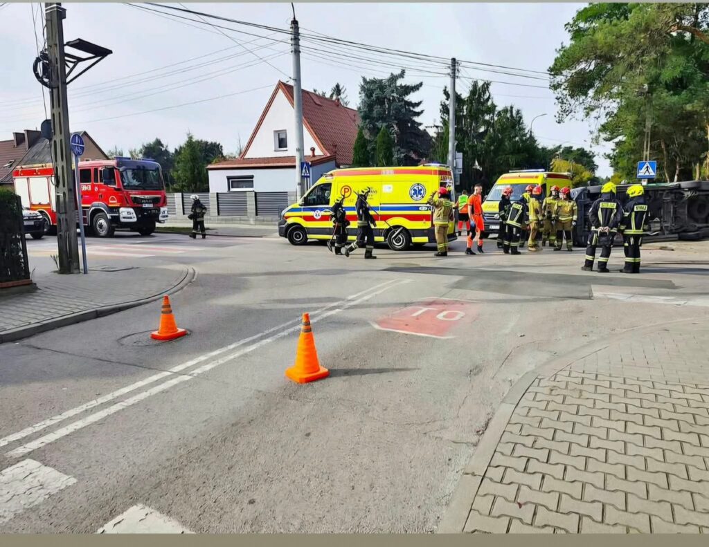 Strażacy i ratownicy medyczni przy dwóch karetkach i wozie strażackim na skrzyżowaniu zabezpieczonym pachołkami.