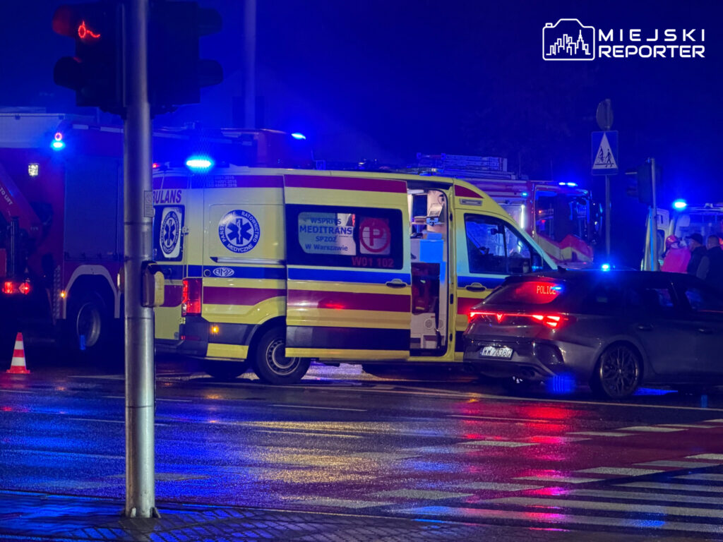 Karetka z otwartymi drzwiami i radiowóz stoją na mokrym skrzyżowaniu, oświetlone niebieskimi sygnałami.