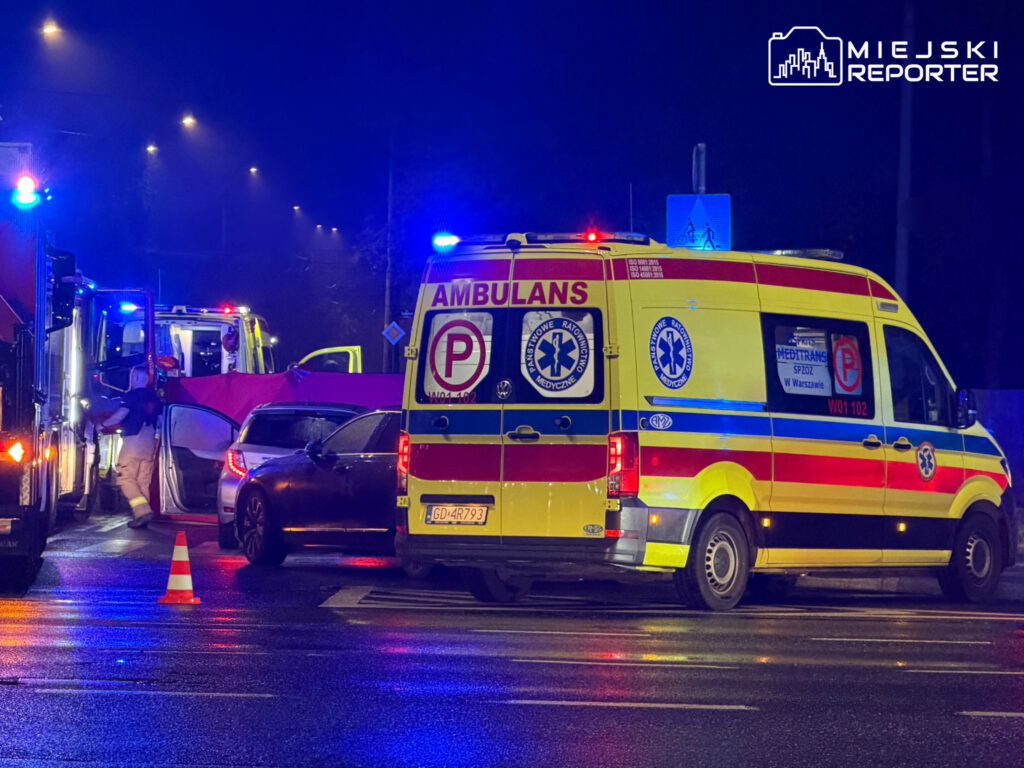 Żółta karetka i wóz strażacki z włączonymi sygnałami świetlnymi stoją nocą na mokrej jezdni obok samochodów.