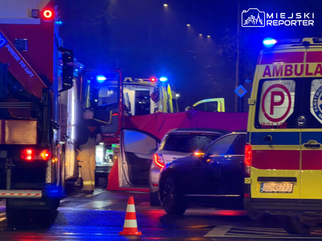 Strażacy i ratownicy medyczni pracują przy samochodach na nocnej, deszczowej ulicy, obok stoją wozy służb ratunkowych.