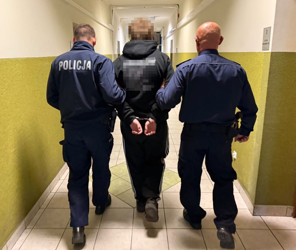 Dwóch policjantów prowadzi skutego kajdankami mężczyznę korytarzem budynku.