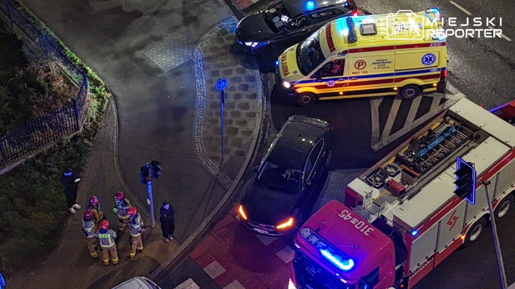 Karetka, radiowóz i wóz strażacki stoją na nocnym skrzyżowaniu, obok którego ratownicy rozmawiają.