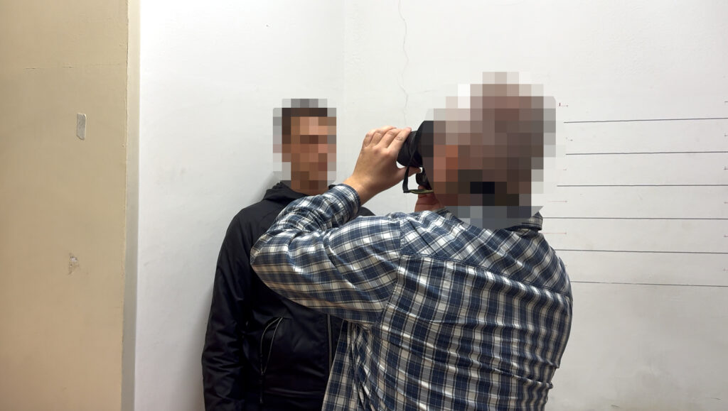 Mężczyzna w kraciastej koszuli fotografuje stojącego przy ścianie mężczyznę na tle podziałki wzrostu.